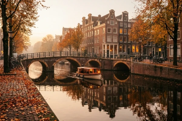 Autumn golden-hour view of an Amsterdam canal with gabled houses, fallen leaves and a lit bridge